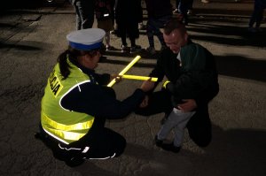 Policjantka z ruchu drogowego zakłada dziecku opaskę odblaskową na rękaw. Obok inni uczestnicy akcji. Zdjęcie wykonane w nocy.