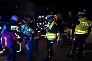 Policjantki z ruchu drogowego podczas akcji profilaktycznej rozdają opaski odblaskowe, zdjęcie wykonane w nocy. Obok policyjny radiowóz.