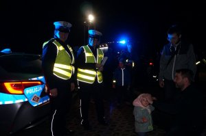 Policjantki z ruchu drogowego podczas akcji profilaktycznej rozdają opaski odblaskowe, zdjęcie wykonane w nocy. Obok policyjny radiowóz.