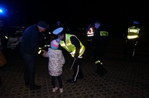 Policjantka z ruchu drogowego podczas akcji profilaktycznej zakłada opaskę odblaskową dziecku, zdjęcie wykonane w nocy. Obok policyjny radiowóz.