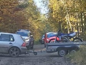 Samochód osobowy z przyczepką, na której jest motocykl crossowy stoi w lesie. Na drugim planie radiowóz terenowy Policji.