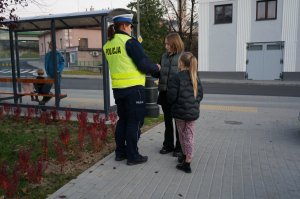 Policjantki rozdają odblaski podczas akcji profilaktycznej. Na drugim planie strażacy ochotnicy.