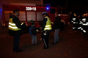 Policjantki rozdają odblaski podczas akcji profilaktycznej. Na drugim planie strażacy ochotnicy.