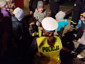 Policjantka z ruchu drogowego zakłada uczestnikowi akcji profilaktycznej opaskę odblaskową. Zdjęcie wykonane w po zmierzchu.