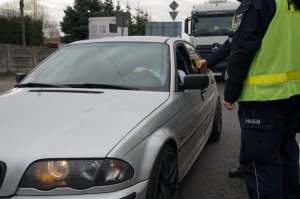 Dwaj policjanci kontrolują stan trzeźwości kierowcy używając ręcznego alkomatu. Zdjęcie wykonane na drodze w dzień.