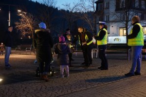 Policjantka w towarzystwie dwóch dziennikarzy rozdaje opaski odblaskowe.