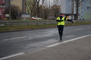 Policjant z drogówki stoi na pasie drogi. Daje znak ręką oraz tarczą drogową, tzw. lizakiem do zjechania na pobocze. Zdjęcie wykonane w pochmurny dzień.