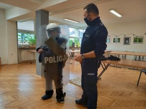 Dzielnicowy prezentuje dziecku policyjne wyposażenie wykorzystywane do zabezpieczania imprez masowych. Ma ono założone ochraniacze, kask, w ręku trzyma tarcze. Zdjęcie wykonane w sali konferencyjnej.