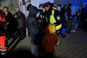 Policjantka rozdaje dzieciom, uczestnikom akcji profilaktycznej opaski odblaskowe. Zdjęcie wykonane po zmierzchu, na dworze.
