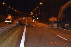 Fotografia z miejsca wypadku drogowego. Na drodze ekspresowej roztrzaskane elementy pojazdu osobowego i maszyna budowlana. Zdjęcie wykonane w nocy.