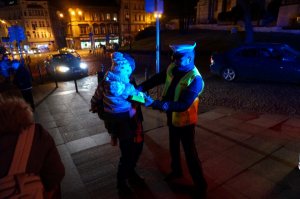 Policjant zakłada opaskę odblaskową dziecku trzymanemu na rękach przez rodzica. Zdjęcie wykonane po zmierzchu.