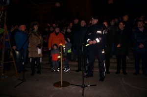 Policjant stoi i wypowiada się do mikrofonu, obok mieszkańcy miasta. Zdjęcie wykonane po zmierzchu.