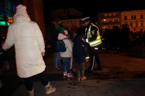 Policjantka zakłada opaskę odblaskową dzieciom podczas akcji profilaktycznej na ulicy. Zdjęcie wykonane po zmierzchu.