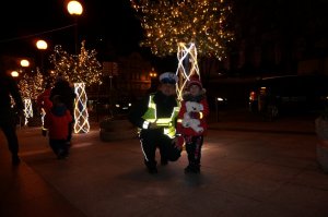 Policjantka kuca koło dziecka i pozują do zdjęcia podczas akcji profilaktycznej na ulicy. Zdjęcie wykonane po zmierzchu.