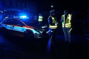 Policjantka udziela wywiadu radiowego. W tle radiowóz. Zdjęcie wykonane na ulicy podczas akcji profilaktycznej po zmierzchu.