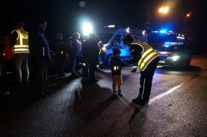 Policjantka pochyla się przy dziecku i zakłada mu opaskę odblaskową. W tle radiowóz i wóz strażacki. Zdjęcie wykonane na ulicy podczas akcji profilaktycznej po zmierzchu.