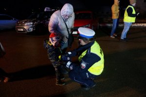 Policjantka kuca przy dziecku i zakłada mu opaskę odblaskową. Zdjęcie wykonane na ulicy podczas akcji profilaktycznej po zmierzchu.