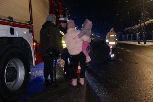 Policjantka rozmawia z dwoma kobieta, z których jedna trzyma dziecko na rękach. Stoją obok wozu strażackiego. Zdjęcie wykonane na ulicy podczas akcji profilaktycznej po zmierzchu.