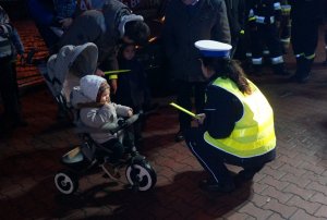 Policjantka z ruchu drogowego kuca przy dziecku. Zdjęcie wykonane na parkingu zewnętrznym podczas akcji profilaktycznej po zmroku.