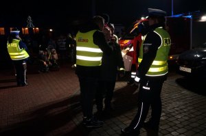 Policjantka ruchu drogowego stoi obok redaktora radiowego, który rozmawia z pieszym. Zdjęcie wykonane na parkingu zewnętrznym podczas akcji profilaktycznej po zmroku.