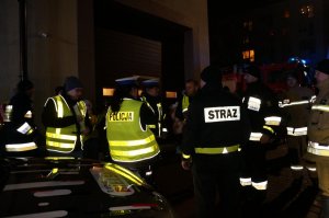 Policjanci i strażacy pożarni rozmawiają przed siedzibą OSP. Zdjęcie wykonane po zmroku.