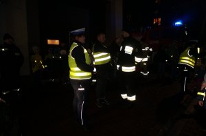 Dziennikarz radiowy prowadzi wywiad ze strażakiem OSP, obok stoi policjant z ruchu drogowego. Zdjęcie wykonane po zmroku.