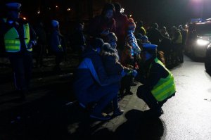 Policjantka z ruchu drogowego kuca przy dziecku. Zdjęcie wykonane na parkingu zewnętrznym podczas akcji profilaktycznej po zmroku.