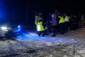 Policjantka z ruchu drogowego kuca przy dziecku. Obok inna policjantka rozdaje opaski odblaskowe innym pieszym. Zdjęcie wykonane na parkingu zewnętrznym podczas akcji profilaktycznej po zmroku.