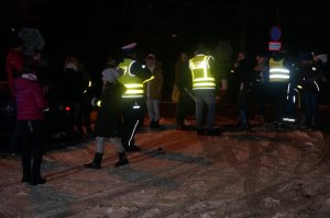 Policjanci i redaktorzy mediów rozdają opaski uczestnikom akcji profilaktycznej. Zdjęcie wykonane po zmroku.