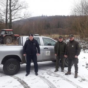 Policjant z dwoma strażnikami leśnymi pozuje do zdjęcia przy samochodzie. Zdjęcie wykonane w okolicy lasu, zimą w dzień.