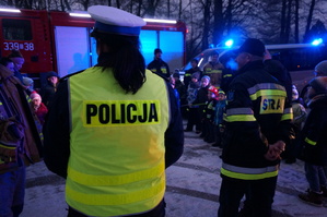 Policjantka i strażak stoją tyłem do robiącego zdjęcie. Na drugim planie wóz strażacki.