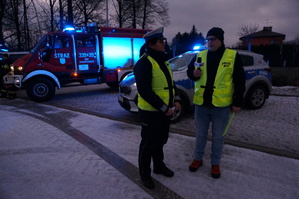 Policjantka ruchu drogowego udziela wywiad radiowego. W tle wozy strażackie i radiowóz policyjny. Zdjęcie wykonane w dzień na parkingu.