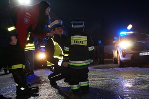 Policjantka ruchu drogowego przyklęka przy dziecku ubranym w strój strażaka. W tle radiowóz policyjny. Zdjęcie wykonane po zmierzchu na parkingu.