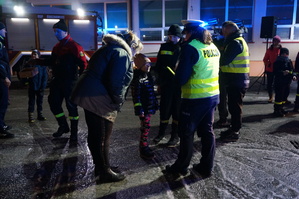 Policjantka z ruchu drogowego rozmawia z dzieckiem. Zdjęcie wykonane podczas akcji profilaktycznej na parkingu, po zmroku. Obok dorosły opiekun dziecka.