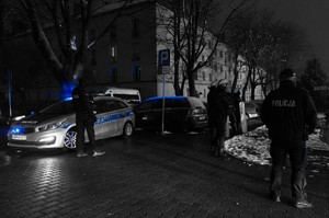 Policjant stoi przy radiowozie, obok inni policjanci stoją przy wejściu na stadion piłkarski. Czarno-białe zdjęcie z niebieskimi efektami.