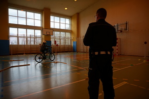 Policjant ruchu drogowego prowadzi test sprawności jazdy rowerem na torze przeszkód ustawionym na sali gimnastycznej.