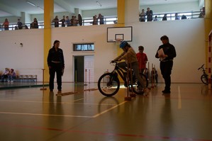 Policjantki z ruchu drogowego prowadzą test sprawności jazdy na rowerze po torze przeszkód ustawionym na sali gimnastycznej.