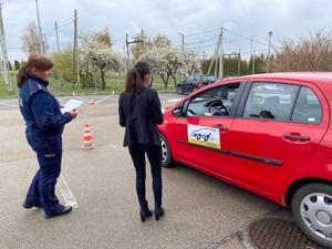 Policjantka obserwuje jadącego czerwonym samochodem osobowym uczestnika konkursu motoryzacyjnego na placu manewrowym.