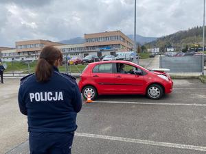 Policjantka obserwuje jadącego czerwonym samochodem osobowym uczestnika konkursu motoryzacyjnego na placu manewrowym.