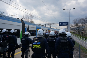 Policjanci z oddziału prewencji stoją obok pociągu, przed nimi stoją kibice