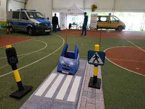 Pluszowy samochodzik do nauki dzieci zasad kodeksu drogowego stoi na specjalnej ścieżce. W tle policyjny radiowóz