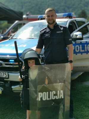 Policjant pozuje do zdjęcia z dzieckiem ubranym w policyjny sprzęt ochronny, w tle terenowy radiowóz