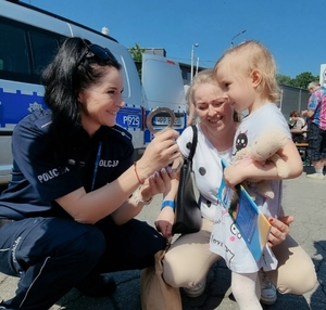 Policjantka kuca przy dziecku i jego opiekunce, pokazuje dziewczynce policyjne kajdanki.