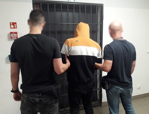 Policjanci w ubiorze cywilnym stoją z zatrzymanym przed wejściem z kratą.