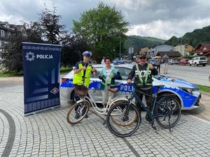 Policjant z ruchu drogowego pozuje do zdjęcia z parą rowerzystów na tle radiowozu.