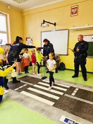 Dwie policjantki i policjant uczą dzieci wlaściwego przechodzenia przez rozlożone przejście dla pieszych.