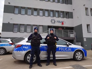 Na zdjęciu umundurowani policjanci stoją przed oznakowanym radiowozem. W tle budynek komendy Policji.