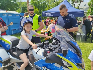 Na motocyklu policyjnym siedzi dziewczynka z założoną policyjną czapka. Obok niej stoi dwóch policjantów.