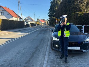 Na obrazku widać policjantkę stojącą po prawej stronie ulicy , przed samochodem mierzącą prędkość za pomocą radaru.