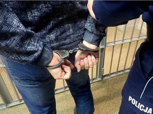 Zdjęcie przedstawia mężczyznę stojącego tyłem z rękami w kajdankach. po jego prawej stronie stoi policjant